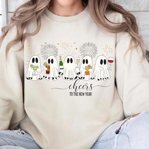 K&ouml;nnte beinhalten: Cremefarbenes Sweatshirt mit festlichem Design. Es zeigt Cartoon-Geister, die Cocktails, Champagner und Wein halten. Der Schriftzug "Cheers to the New Year" steht darunter, mit Feuerwerk im Hintergrund.
