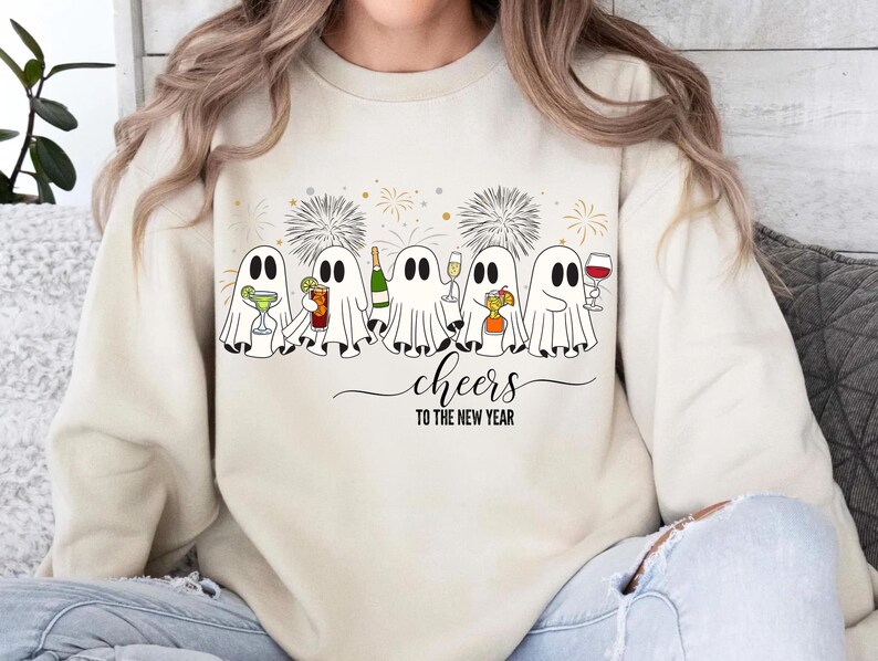K&ouml;nnte beinhalten: Cremefarbenes Sweatshirt mit festlichem Design. Es zeigt Cartoon-Geister, die Cocktails, Champagner und Wein halten. Der Schriftzug "Cheers to the New Year" steht darunter, mit Feuerwerk im Hintergrund.