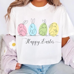 May include: White t-shirt with a graphic of four line-drawn bunnies in different colors, each sitting inside an egg. The text "Happy Easter" is written below the bunnies.