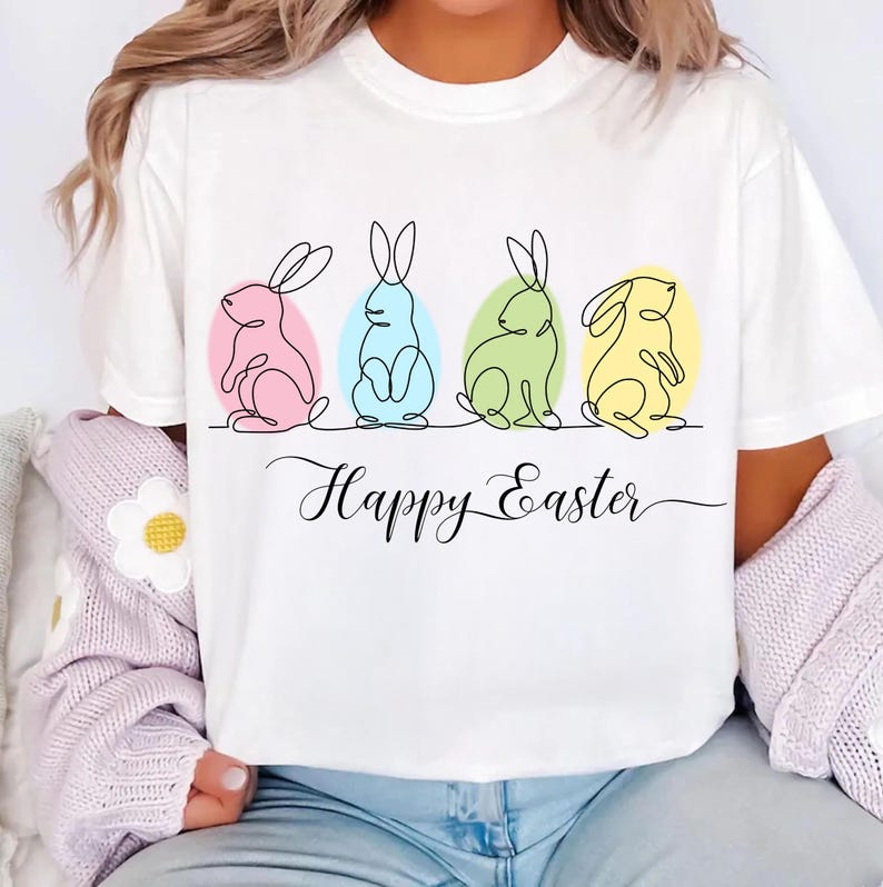 May include: White t-shirt with a graphic of four line-drawn bunnies in different colors, each sitting inside an egg. The text "Happy Easter" is written below the bunnies.