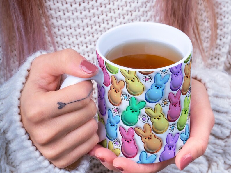 May include: A white ceramic mug with a colorful pattern of pastel-colored bunny shapes and white daisies. The mug is filled with a light brown liquid.