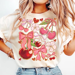 May include: A white t-shirt with a pink and red cherry and bow design. The design features cherries, bows, flowers, and hearts.