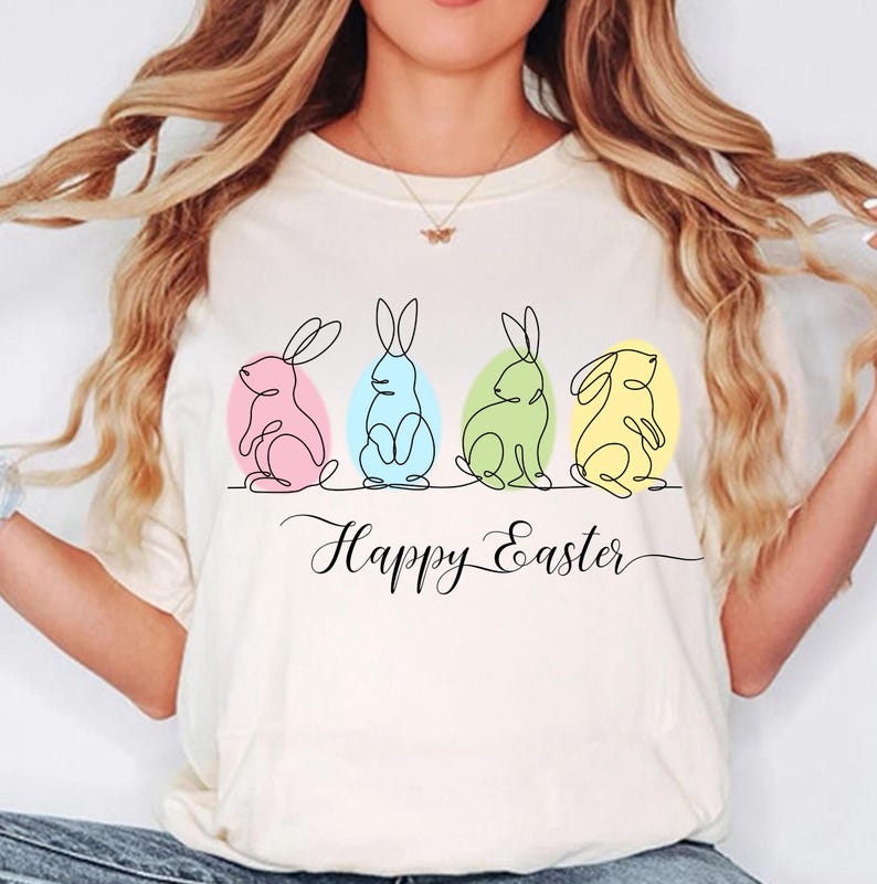 May include: White t-shirt with a graphic of four Easter bunnies in a line, each bunny is a different color and inside an egg shape. The text "Happy Easter" is written below the bunnies.