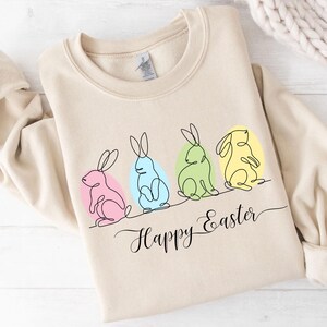 May include: A beige sweatshirt with a graphic of four line-drawn bunnies in pastel colors, sitting in a row. The bunnies are surrounded by pastel-colored circles. The text "Happy Easter" is written in cursive below the bunnies.