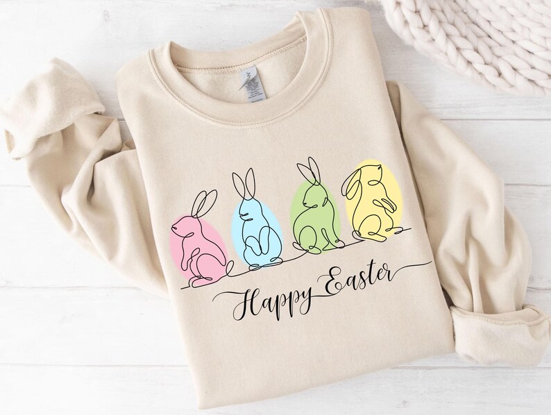 May include: A beige sweatshirt with a graphic of four line-drawn bunnies in pastel colors, sitting in a row. The bunnies are surrounded by pastel-colored circles. The text "Happy Easter" is written in cursive below the bunnies.
