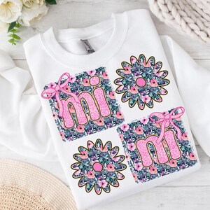 May include: White sweatshirt with a pink "mi" and "mi" design on a floral background. The letters are outlined in gold and have a pink bow on each.