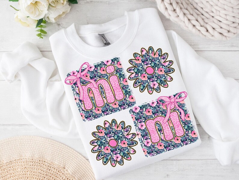 May include: White sweatshirt with a pink "mi" and "mi" design on a floral background. The letters are outlined in gold and have a pink bow on each.