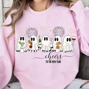 K&ouml;nnte beinhalten: Hellrosa Sweatshirt mit einem Motiv von f&uuml;nf Cartoon-Geistern, die mit Getr&auml;nken ansto&szlig;en. Der Schriftzug "cheers to the new year" steht darunter. Feuerwerk im Hintergrund rundet das festliche Design ab.