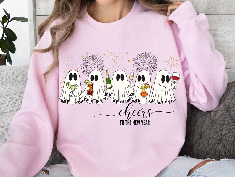 K&ouml;nnte beinhalten: Hellrosa Sweatshirt mit einem Motiv von f&uuml;nf Cartoon-Geistern, die mit Getr&auml;nken ansto&szlig;en. Der Schriftzug "cheers to the new year" steht darunter. Feuerwerk im Hintergrund rundet das festliche Design ab.