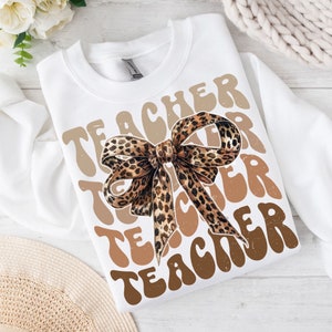 Puede incluir: Sudadera blanca con la palabra "TEACHER" repetida en marrón y beige en una fuente retro. Un lazo de estampado de leopardo marrón y negro está centrado en la sudadera.