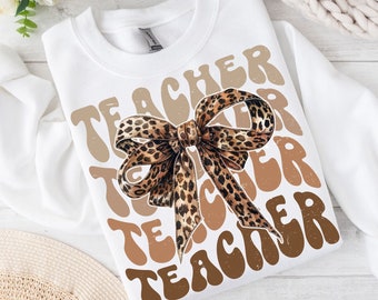 May include: White sweatshirt with the word "TEACHER" repeated in brown and tan colors in a retro font. A brown and black leopard print bow is centered on the shirt.
