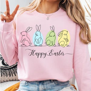 May include: Pink sweatshirt with a graphic of four line-drawn bunnies in pastel colors, each with an egg behind it. The text "Happy Easter" is written below the bunnies.