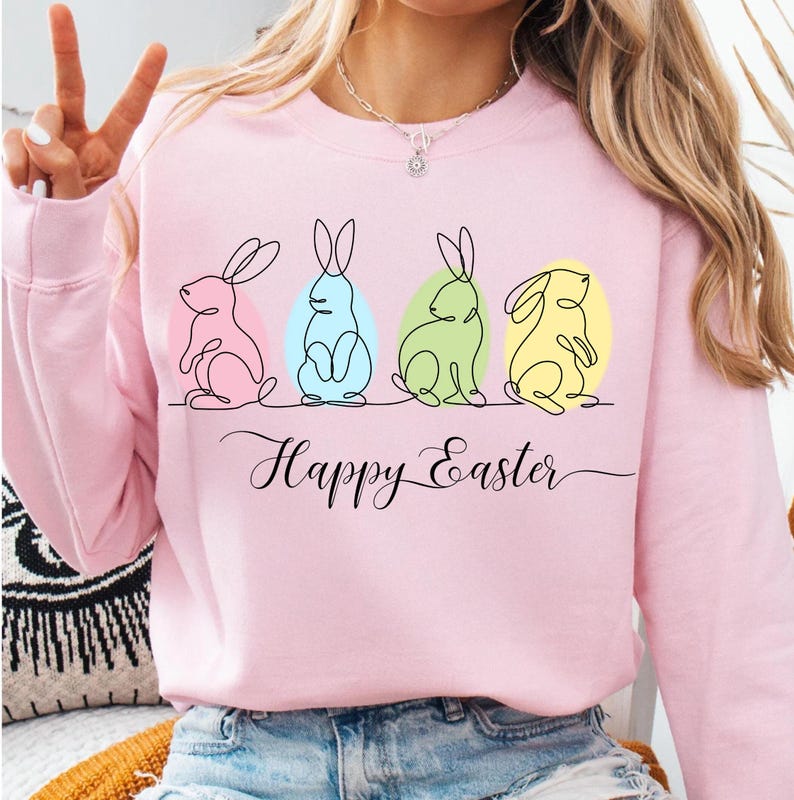 May include: Pink sweatshirt with a graphic of four line-drawn bunnies in pastel colors, each with an egg behind it. The text "Happy Easter" is written below the bunnies.