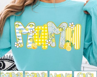 May include: A turquoise sweatshirt featuring the word "MAMA" in a decorative font. The letters are yellow and adorned with lemons, stripes, and floral designs. White daisies are placed around the letters.