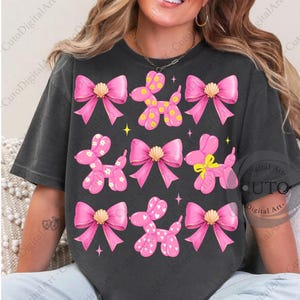 May include: Dark gray t-shirt featuring a repeating pattern of pink balloon animals and bows. The balloon animals have polka dots, hearts, and flower designs. The bows have gold seashell accents. The design includes yellow stars.