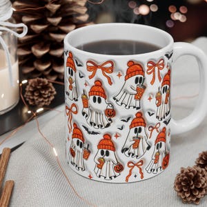 May include: White ceramic mug featuring a repeating pattern of cartoon ghosts wearing orange beanies and holding coffee cups and jack-o'-lanterns. The mug has a white handle and is filled with a dark liquid. Halloween theme.