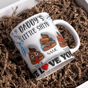 K&ouml;nnte beinhalten: Wei&szlig;e Keramiktasse mit einem lustigen Spruch "Daddy's Little Shits" und Kack-Emoji-Grafiken. Die Tasse hat zwei leere Stellen f&uuml;r Namen und den Text "We Love You".
