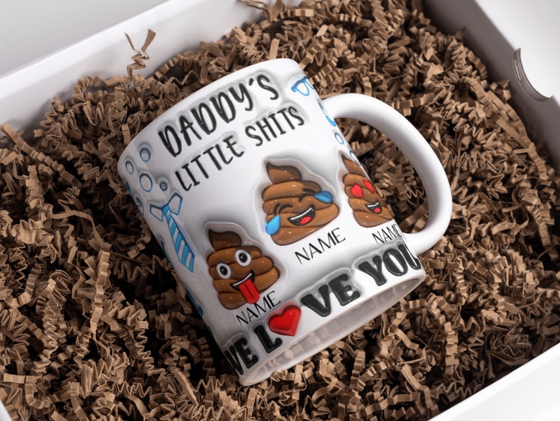 K&ouml;nnte beinhalten: Wei&szlig;e Keramiktasse mit einem lustigen Spruch "Daddy's Little Shits" und Kack-Emoji-Grafiken. Die Tasse hat zwei leere Stellen f&uuml;r Namen und den Text "We Love You".