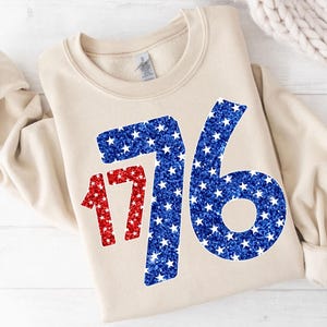 May include: A beige sweatshirt with a large red and blue glitter design of the number 1776. The numbers are made up of small white stars.