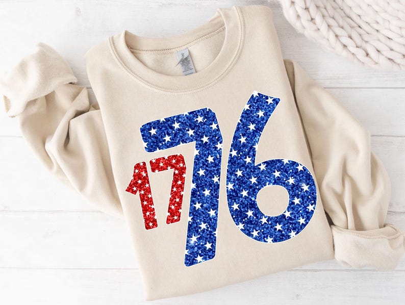 May include: A beige sweatshirt with a large red and blue glitter design of the number 1776. The numbers are made up of small white stars.