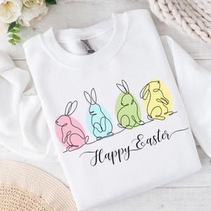 May include: White sweatshirt with a line drawing of four bunnies in different colors, sitting on eggs. The text "Happy Easter" is written below the bunnies.
