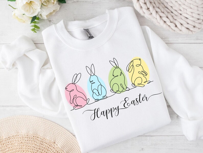 May include: White sweatshirt with a line drawing of four bunnies in different colors, sitting on eggs. The text "Happy Easter" is written below the bunnies.