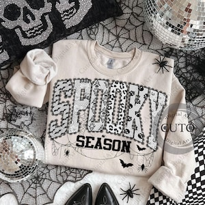 May include: A beige sweatshirt with the words "SPOOKY SEASON" in a black and white design. The word "SPOOKY" is decorated with bats, skulls, and lace. The sweatshirt is surrounded by Halloween-themed decorations, including spiders and a disco ball.