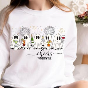 K&ouml;nnte beinhalten: Wei&szlig;es Sweatshirt mit festlichem Design. Cartoon-Geister sind abgebildet, die Cocktails, Champagner und Wein halten. Das Design enth&auml;lt auch Feuerwerk und den Satz "cheers to the new year". Eine lustige Grafik f&uuml;r die Ferienzeit.