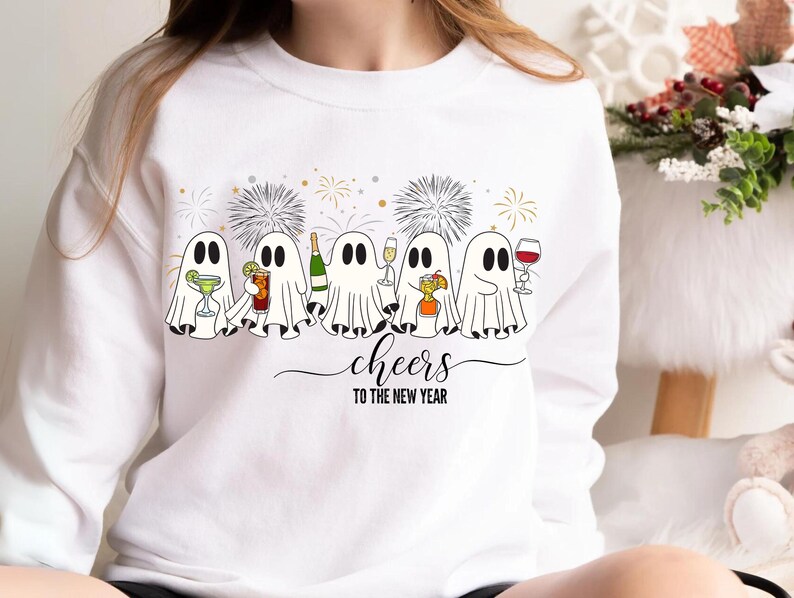 K&ouml;nnte beinhalten: Wei&szlig;es Sweatshirt mit festlichem Design. Cartoon-Geister sind abgebildet, die Cocktails, Champagner und Wein halten. Das Design enth&auml;lt auch Feuerwerk und den Satz "cheers to the new year". Eine lustige Grafik f&uuml;r die Ferienzeit.