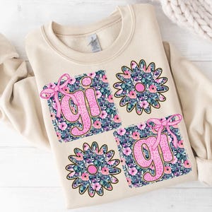 May include: A beige sweatshirt with a floral design featuring the word "girl" in pink letters. The design is made up of four squares with floral patterns and pink bows.