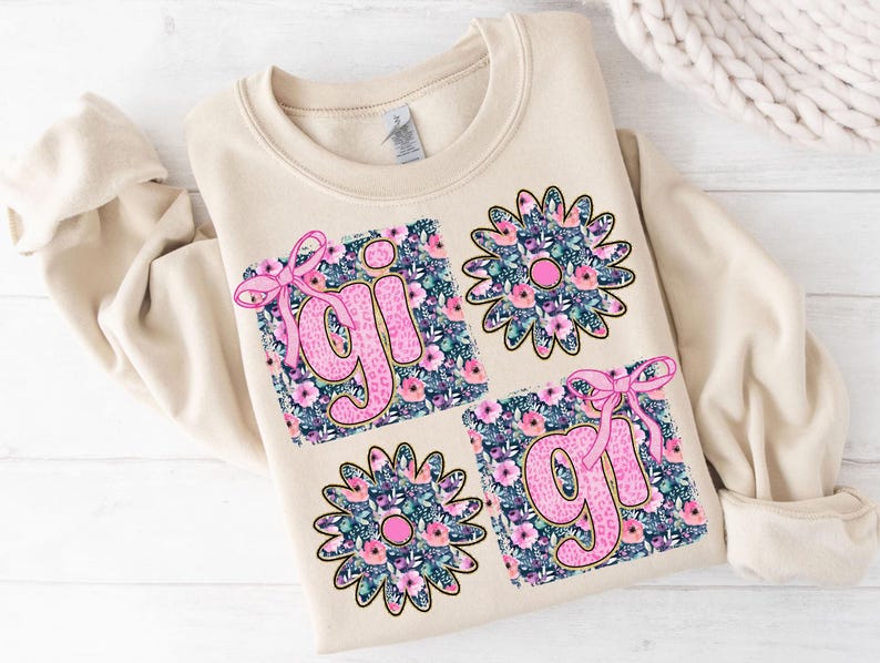 May include: A beige sweatshirt with a floral design featuring the word "girl" in pink letters. The design is made up of four squares with floral patterns and pink bows.