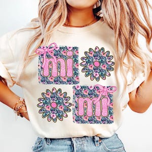May include: A white t-shirt with a floral design and the word "mi" repeated twice. The floral design is in shades of pink, blue, and green.