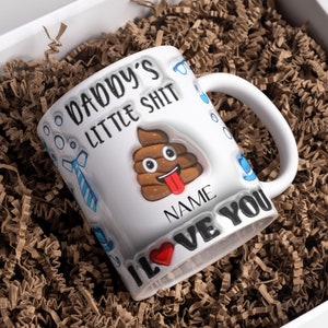 K&ouml;nnte beinhalten: Wei&szlig;e Keramiktasse mit einem braunen Kack-Emoji und dem Text "Daddy's Little Shit" und "I Love You".