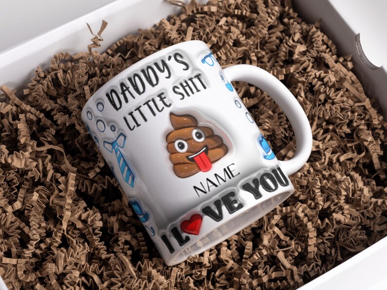 K&ouml;nnte beinhalten: Wei&szlig;e Keramiktasse mit einem braunen Kack-Emoji und dem Text "Daddy's Little Shit" und "I Love You".