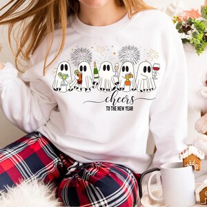 K&ouml;nnte beinhalten: Wei&szlig;es Sweatshirt mit einer Grafik von Cartoon-Geistern, die mit Getr&auml;nken ansto&szlig;en, Feuerwerk und dem Text "cheers to the new year". Die Geister halten Cocktails, Champagner und Wein. Das Sweatshirt wird mit einer rot-blauen karierten Pyjamahose getragen.