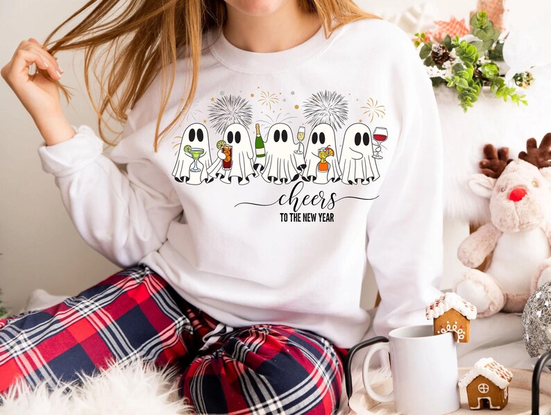 K&ouml;nnte beinhalten: Wei&szlig;es Sweatshirt mit einer Grafik von Cartoon-Geistern, die mit Getr&auml;nken ansto&szlig;en, Feuerwerk und dem Text "cheers to the new year". Die Geister halten Cocktails, Champagner und Wein. Das Sweatshirt wird mit einer rot-blauen karierten Pyjamahose getragen.