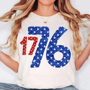 May include: White t-shirt with a red and blue glitter design of the number 1776. The numbers are made up of small white stars on a blue background with red outlines.