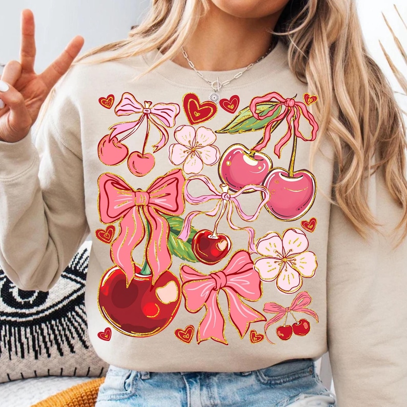 May include: A beige sweatshirt with a pink and red cherry and bow print. The print features cherries, bows, flowers, and hearts.