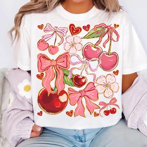 May include: White t-shirt with a pink and gold cherry and bow print. The design features cherries, bows, hearts, and flowers.