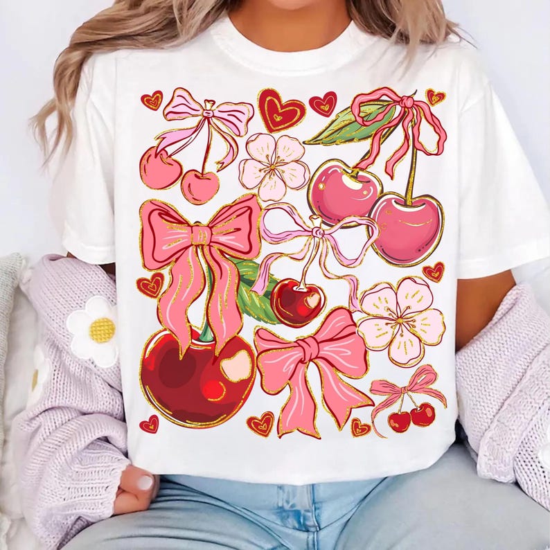 May include: White t-shirt with a pink and gold cherry and bow print. The design features cherries, bows, hearts, and flowers.
