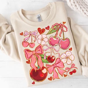 May include: A beige sweatshirt with a graphic print of pink cherries, bows, and flowers. The cherries have green stems and leaves.