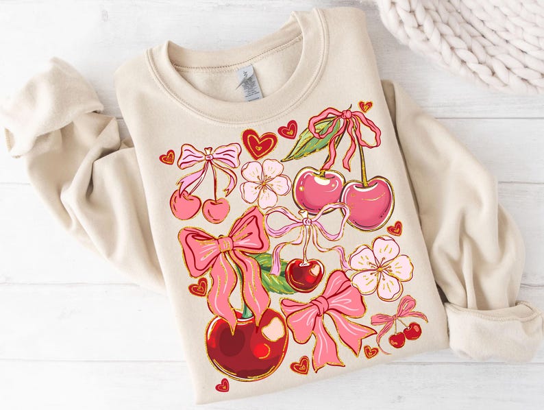 May include: A beige sweatshirt with a graphic print of pink cherries, bows, and flowers. The cherries have green stems and leaves.