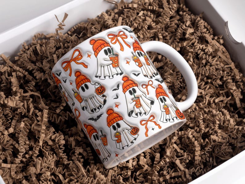 May include: White ceramic mug with a Halloween ghost pattern. The ghosts wear orange beanies and hold drinks and jack-o'-lanterns. The mug is in a white box filled with brown crinkle paper.