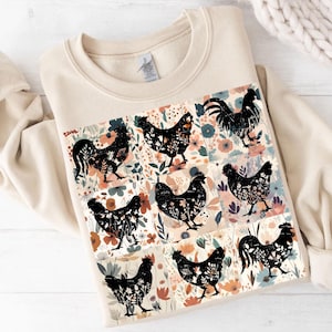 May include: A cream-colored sweatshirt featuring a colorful floral and chicken design. The design includes black chicken silhouettes with floral patterns, set against a backdrop of flowers in various colors. The sweatshirt has a crew neck and long sleeves.
