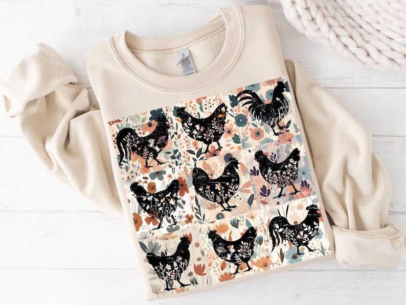 May include: A cream-colored sweatshirt featuring a colorful floral and chicken design. The design includes black chicken silhouettes with floral patterns, set against a backdrop of flowers in various colors. The sweatshirt has a crew neck and long sleeves.