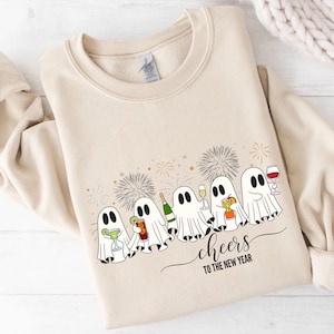 K&ouml;nnte beinhalten: Beiges Sweatshirt mit einer Grafik von Cartoon-Geistern, die mit Getr&auml;nken ansto&szlig;en. Mit Feuerwerk und dem Text "cheers to the new year". Das Sweatshirt hat lange &Auml;rmel und einen Rundhalsausschnitt.