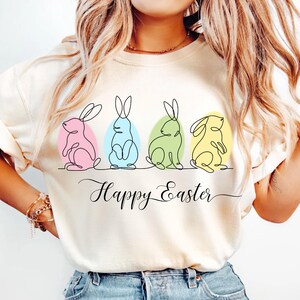 May include: A white t-shirt with a graphic of four Easter bunnies in a line, each bunny is a different color. The bunnies are drawn in a single line style. The text "Happy Easter" is written below the bunnies.