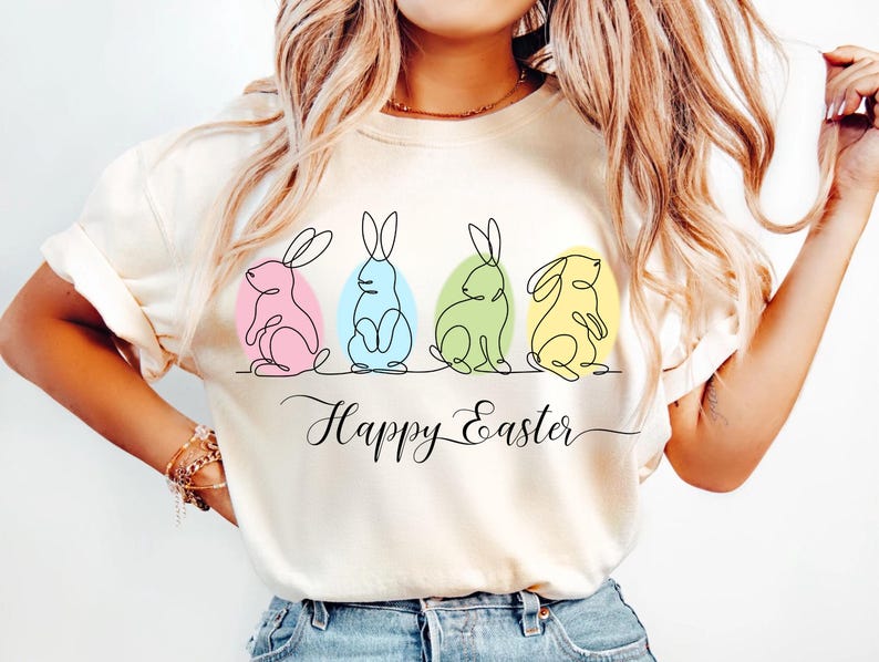 May include: A white t-shirt with a graphic of four Easter bunnies in a line, each bunny is a different color. The bunnies are drawn in a single line style. The text "Happy Easter" is written below the bunnies.