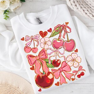 May include: White sweatshirt with a pink and red cherry design. The cherries have pink bows and are surrounded by pink flowers and hearts.