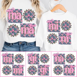 May include: Two white shirts with pink floral designs and the words "mama" and "mini" on them. The shirts are shown on a white background.